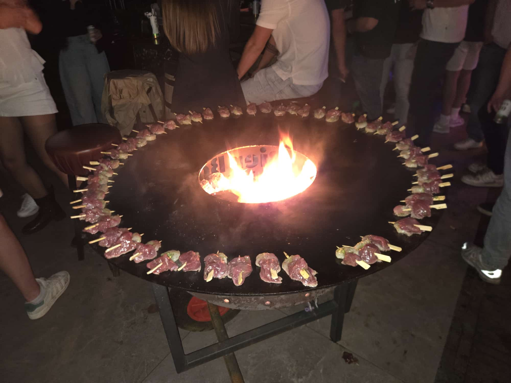 Sfeervolle vuurschaal-barbecue met satéspiesjes en gasten eromheen tijdens een feest